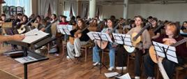 Na zdjęciu uczestnicy III festiwalu Gitarowy Lublin w czasie próby orkiestry gitarowej prowadzonej przez Tomasza Radziszewskiego - Martin - duża grupa około 50 osób. Wszyscy trzymają gitary, przed nimi stoją pulpity. W pierwszym rzędzie siedzi uczennica naszej szkoły Amelia Lewkowicz. Przed nimi widać pulpit i rękę dyrygenta. 
