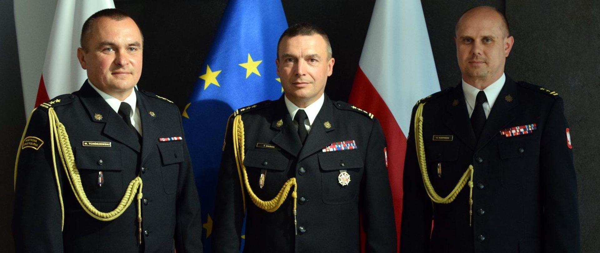 Zdjęcie zrobione wewnątrz pomieszczenia. Na zdjęciu trzech oficerów Państwowej Straży Pożarnej. Za nimi stoją biało-czerwone flagi oraz flaga UE.