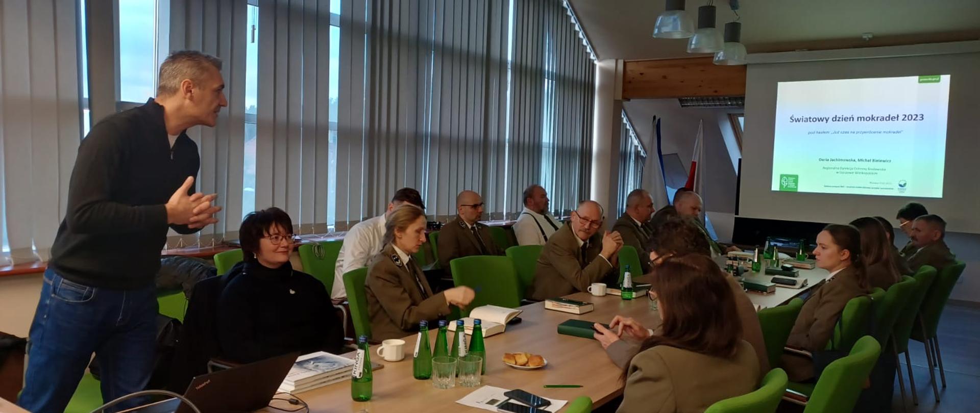 Na drugim planie znajduje się wiszący na ścianie ekran z wyświetlonym zdjęciem. W dużej sali na krzesłach siedzą kobiety i mężczyźni ubrani w mundury leśników.