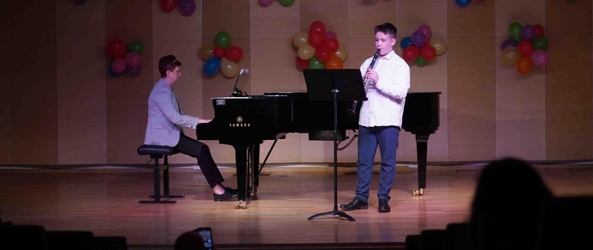 zdjęcie przedstawia ucznia grającego na klarnecie, przed nim pulpit, obok akompaniatorka gra na fortepianie, w tle dekoracja z kolorowych balonów
