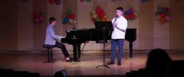 zdjęcie przedstawia ucznia grającego na klarnecie, przed nim pulpit, obok akompaniatorka gra na fortepianie, w tle dekoracja z kolorowych balonów