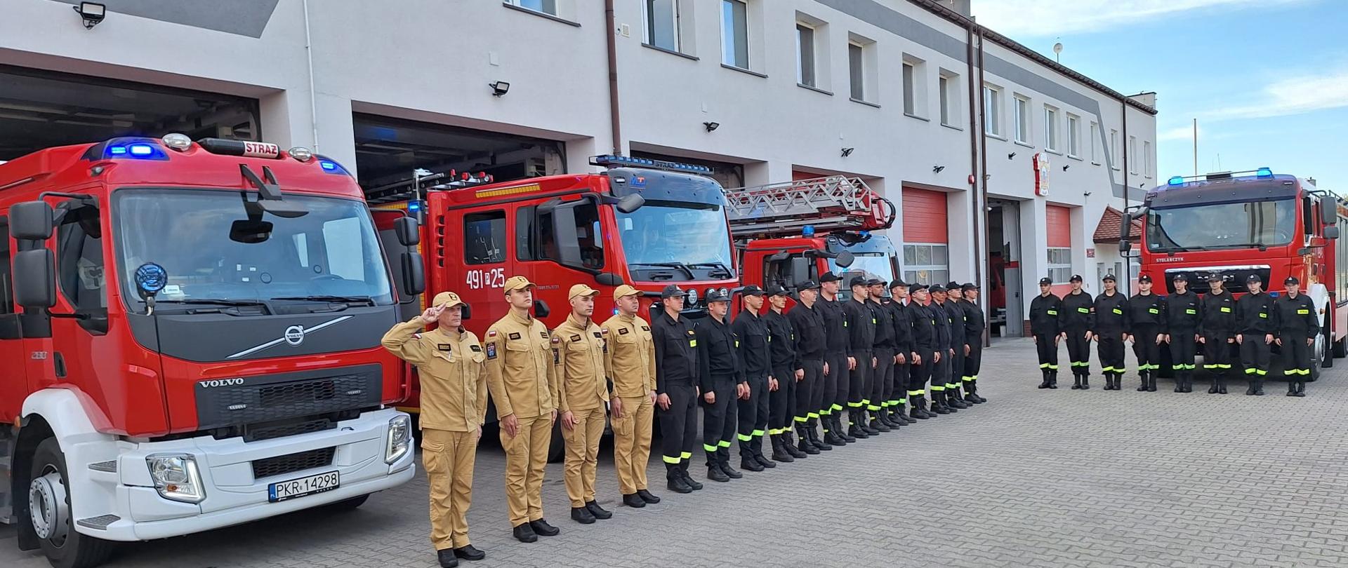 Zdjęcie przedstawia strażaków oddających honor strażakowi, który zginął tragicznie podczas pożaru. Strażacy stoją przed budynkiem JRG na tle samochodów pożarniczych.