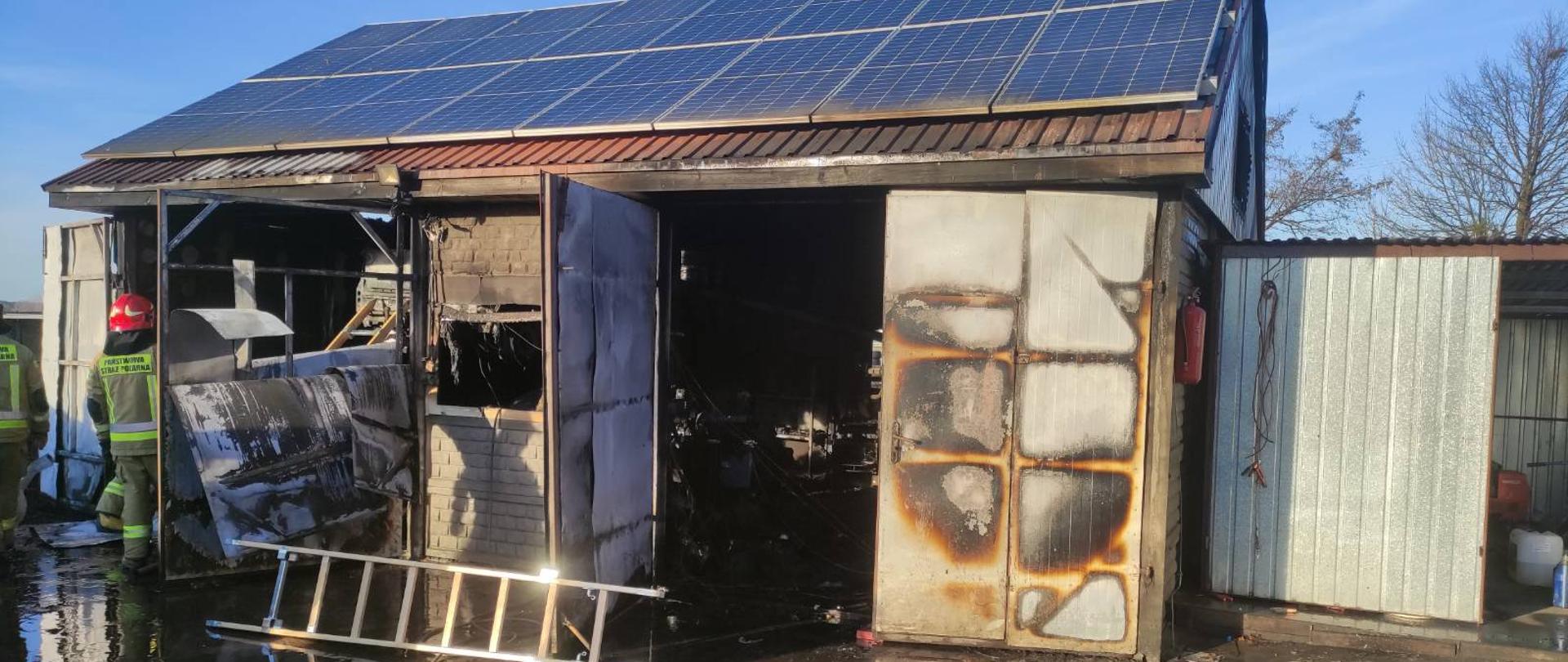 Zdjęcie przedstawia budynek warsztatu samochodowego. Na dachu budynku znajdują się panele fotowoltaiczne. Wrota od warsztatu są otwarte. Wewnątrz wszystko uległo spaleniu. Na zdjęciu widać również strażaków, linię gaśniczą oraz drabinę. 