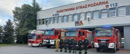 Strażacy stoją w szeregu przed frontem samochodów pożarniczych i JRG Opatów