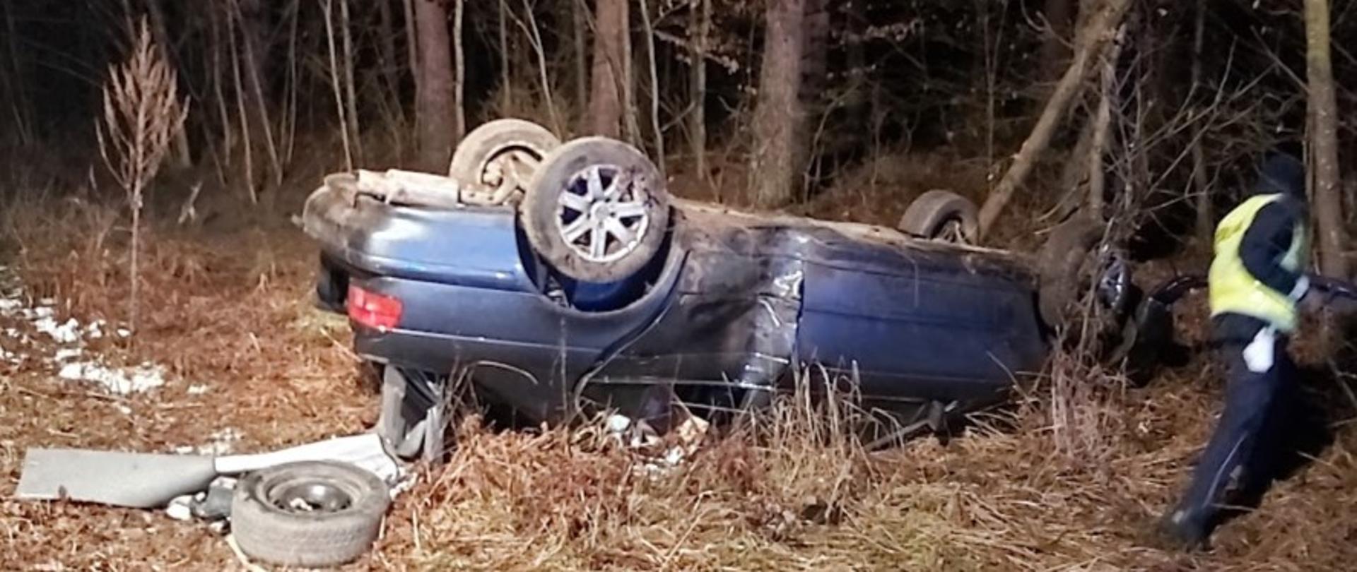 Auto osobowe leży na dachu na poboczu jezdni. W aucie widoczny uszkodzenie tylnych drzwi po uderzeniu w drzewo.