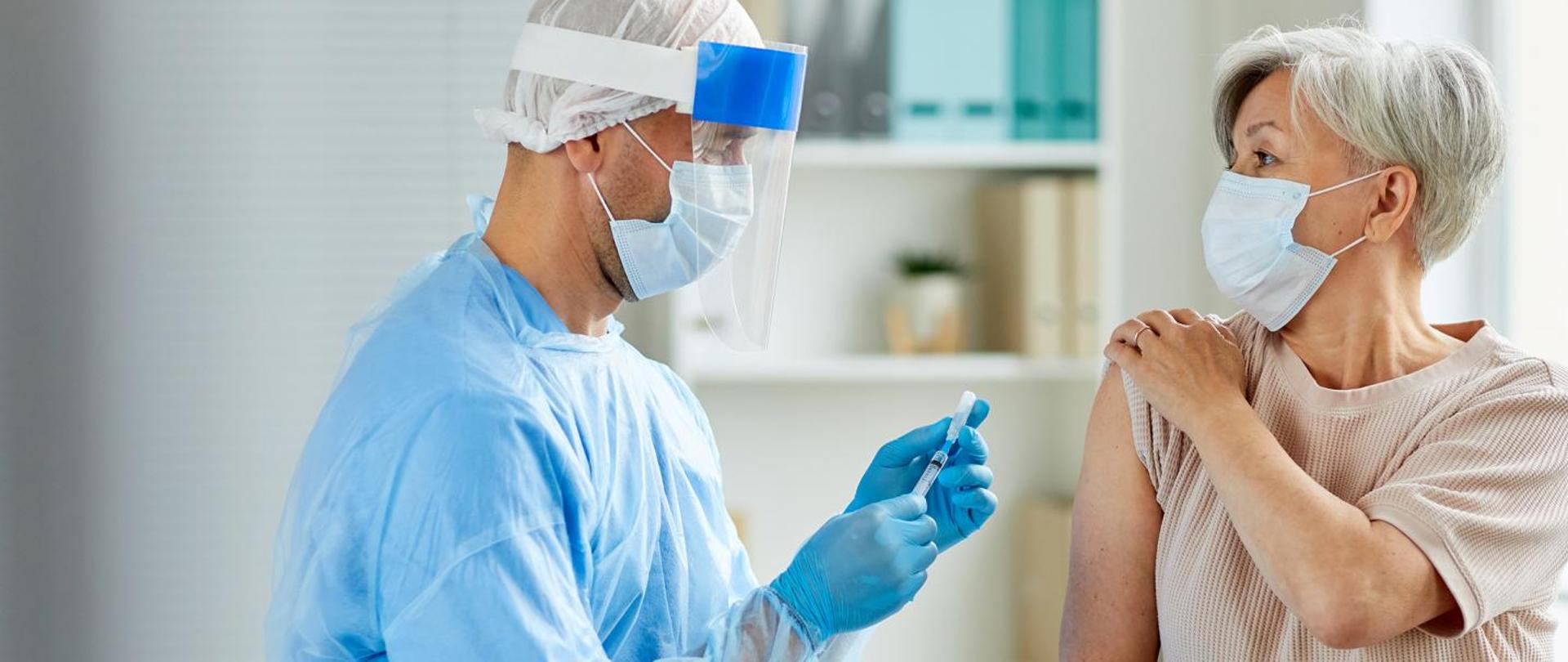 Na zdjęciu znajduje się medyk trzymający w dłoniach strzykawkę. Obok jest pacjentka, która ma odsłonięte ramię. W tle widać wnętrze gabinetu lekarskiego.