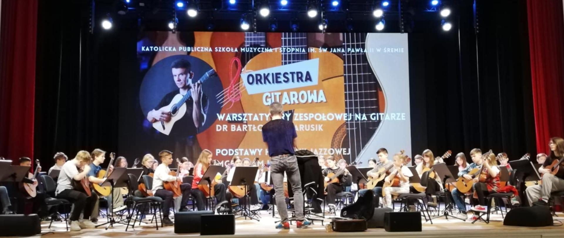 sala koncertowa, na podwyższonej scenie siedzi duża orkiestra gitarowa, za nią duży baner i bordowe kotary