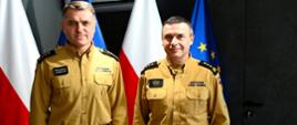 Kolorowa fotografia wykonana w pomieszczeniu. Przedstawia dwóch strażaków w strojach służbowych w pomieszczeniu podkarpackiego komendanta wojewódzkiego PSP po wręczeniu aktu powołania na stanowisko zastępcy komendanta powiatowego PSP w Brzozowie. W tle flagi Polski i Unii Europejskiej.