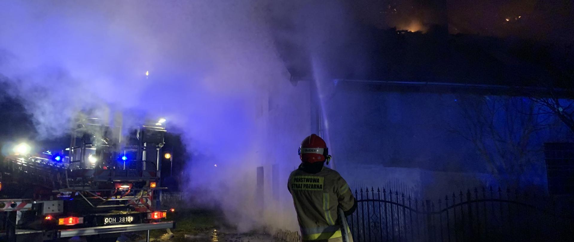 Na zdjęciu widoczny pożar dachu budynku. Pożarem objęta jest górna część dachu dwuspadzistego pokrytego dachówką koloru brązowego. Z dachu wydobywa się duża ilość dymu koloru szarego.
Na pierwszym planie w centralnym miejscu strażak w ubraniu specjalnym koloru piaskowego z czerwonym hełmem na głowie trzyma prądownicę i wąż pożarniczy koloru białego i podaje prąd zwarty wody na dach budynku. Strażak skierowany twarzą do budynku (plecami do fotografującego). Po prawej stronie strażaka widoczna część czarnego ogrodzenia budynku. Po lewej stronie widoczny kawałek podnośnika hydraulicznego z JRG Krapkowice (widoczny tył pojazdu wraz z koszem ratowniczym) rozłożonego na asfaltowej drodze.