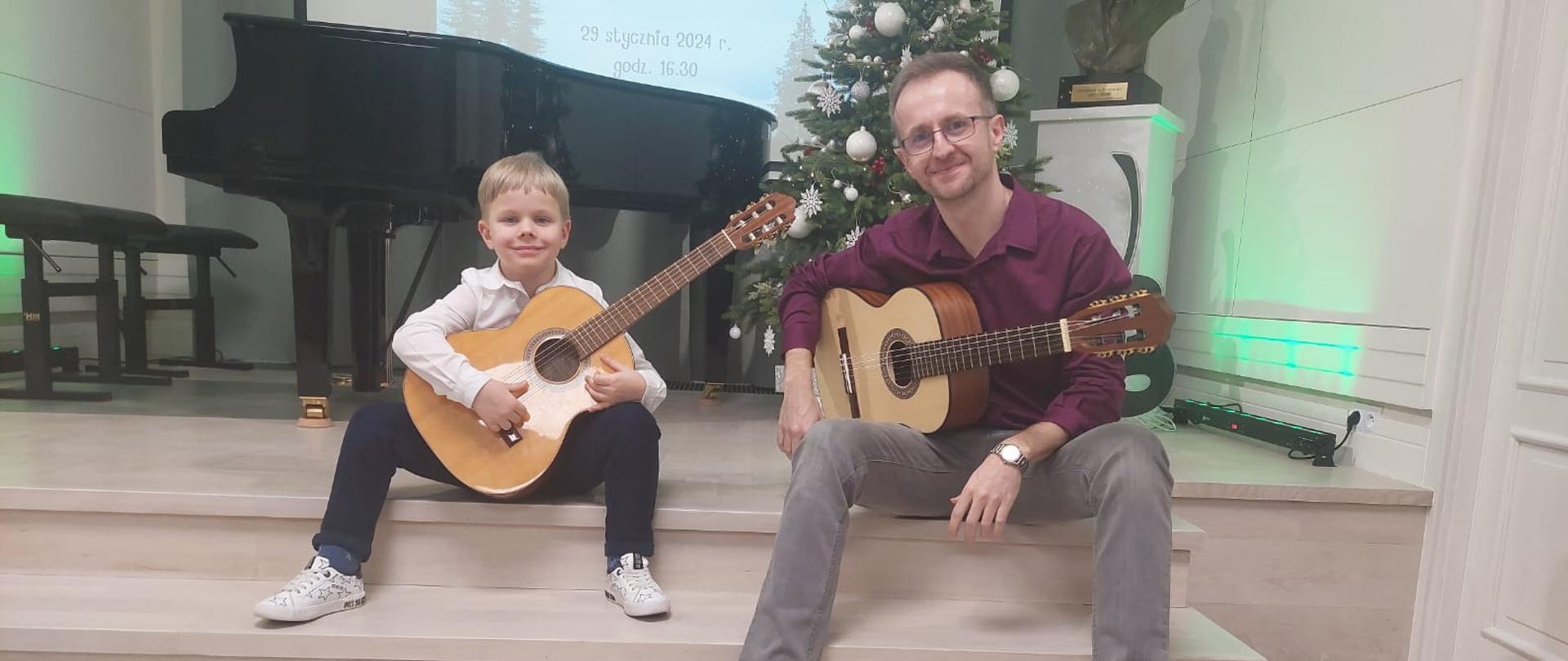 Uczeń i nauczyciel siedzą na stopniach sceny i trzymają na kolanach gitary. Uczeń ma na sobie białą koszulę i czarne spodnie, nauczyciel bordową koszulę i szare spodnie. Z tyłu po prawej stoi ubrana choinka a po lewej fortepian. Scena jest doświetlona na zielono. 