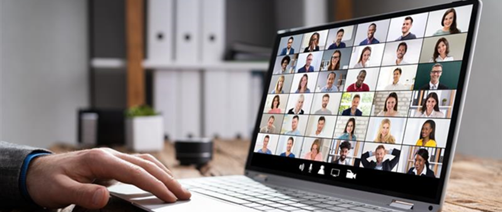 Otwarty laptop, na ekranie kilkadziesiąt okien z wizerunkami osób biorących udział w spotkaniu online. Na klawiaturze męska dłoń. W tle szafka z segregatorami
