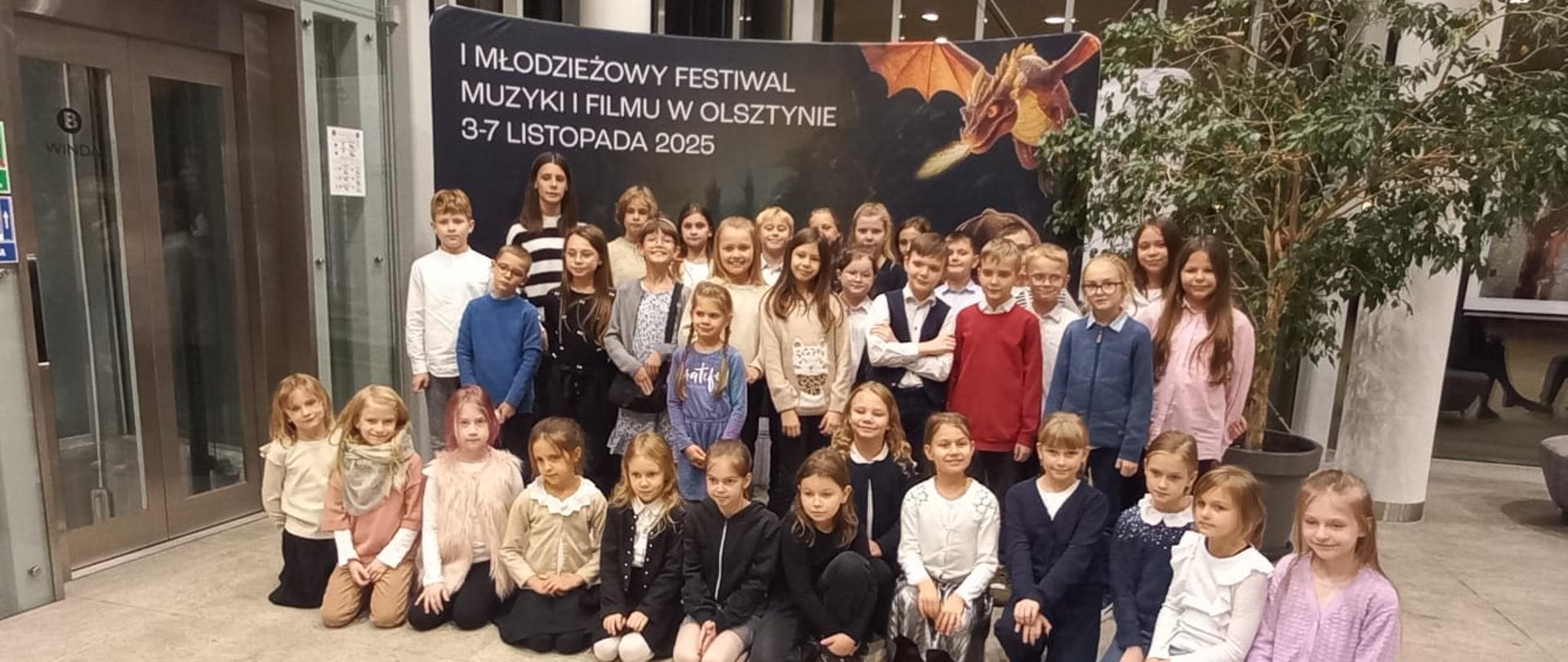 Zdjęcie przedstawia grupę uczniów szkoły ustawioną przed banerem z napisem: "I młodzieżowy festiwal muzyki i filmu w Olsztynie 3-7 listopada 2025"