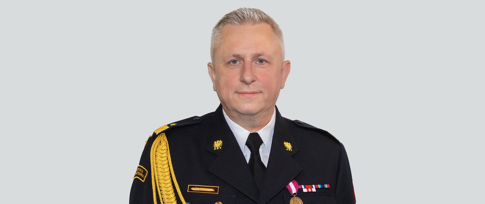 Portretowe zdjęcie funkcjonariusza Państwowej Straży Pożarnej w czarnym mundurze galowym, wykonane na jasnym, jednolitym tle. Mężczyzna stoi przodem do obiektywu, patrzy wprost, ma krótkie siwe włosy i spokojny wyraz twarzy. Na mundurze widoczne są złote dystynkcje, odznaki, baretkowe odznaczenia, medal oraz złoty sznur galowy przewieszony przez prawe ramię i pierś. Na lewej piersi znajduje się imienna plakietka z nazwiskiem „Sierpatowski”.