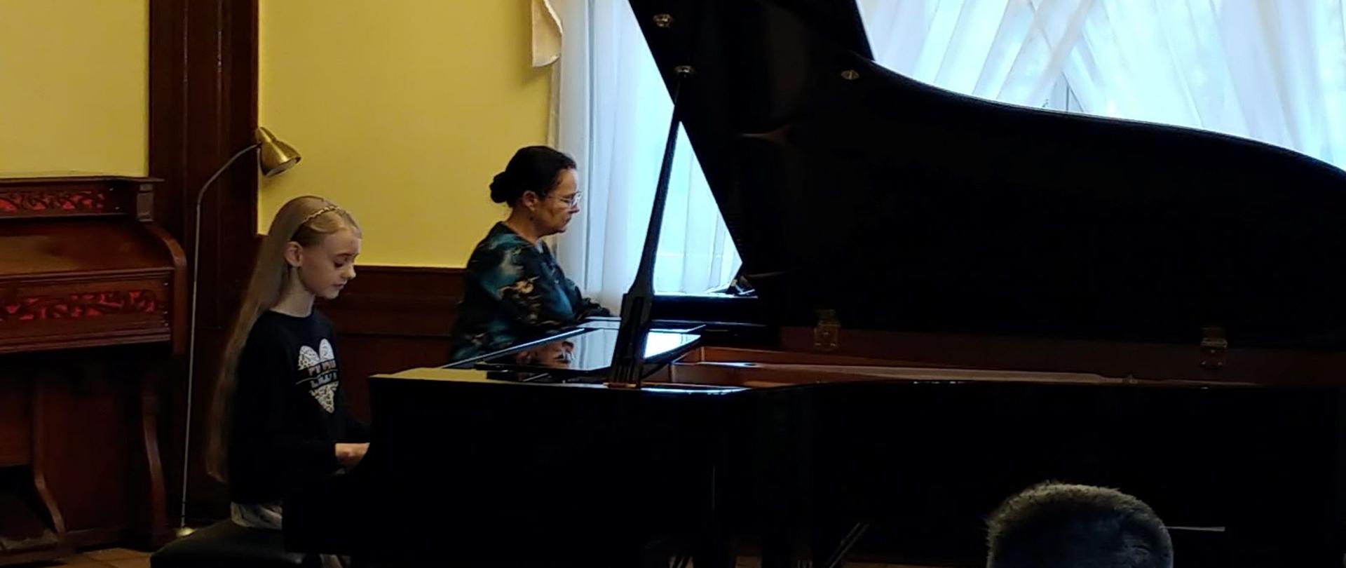 Uczennica oraz Jaj nauczycielka siedzą przy fortepianach. Prezentują utwór muzyczny podczas koncertu Sekcji Fortepianu.