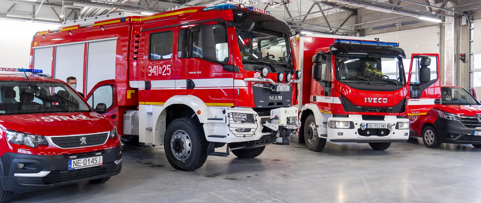 Cztery pojazdy pożarnicze widziane od przodu, zaparkowane w garażu strażackiej remizy.