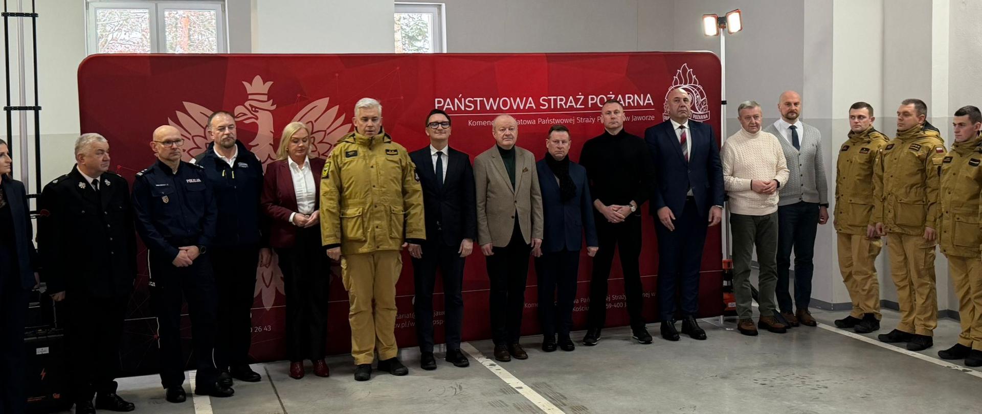 Grupa osób stoi w szeregu na tle czerwonej ścianki z emblematem straży pożarnej i napisem o komendzie powiatowej w Jaworze. Wśród nich są funkcjonariusze w mundurach straży i policji oraz osoby w eleganckich strojach. Scena ma charakter oficjalny i wygląda na uroczyste wydarzenie służbowe.