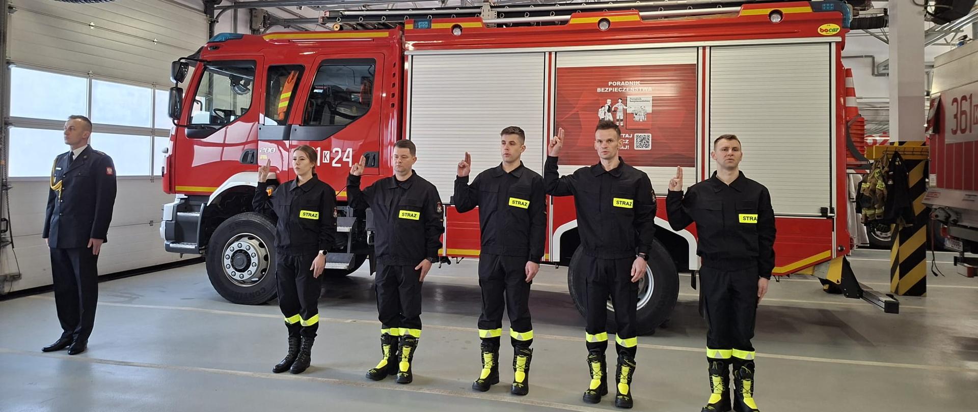 Na garażu obok pożarniczego samochodu bojowego w rzędzie stoją strażacy ubrani w mundury koszarowe. Unoszą prawe dłonie do ślubowania