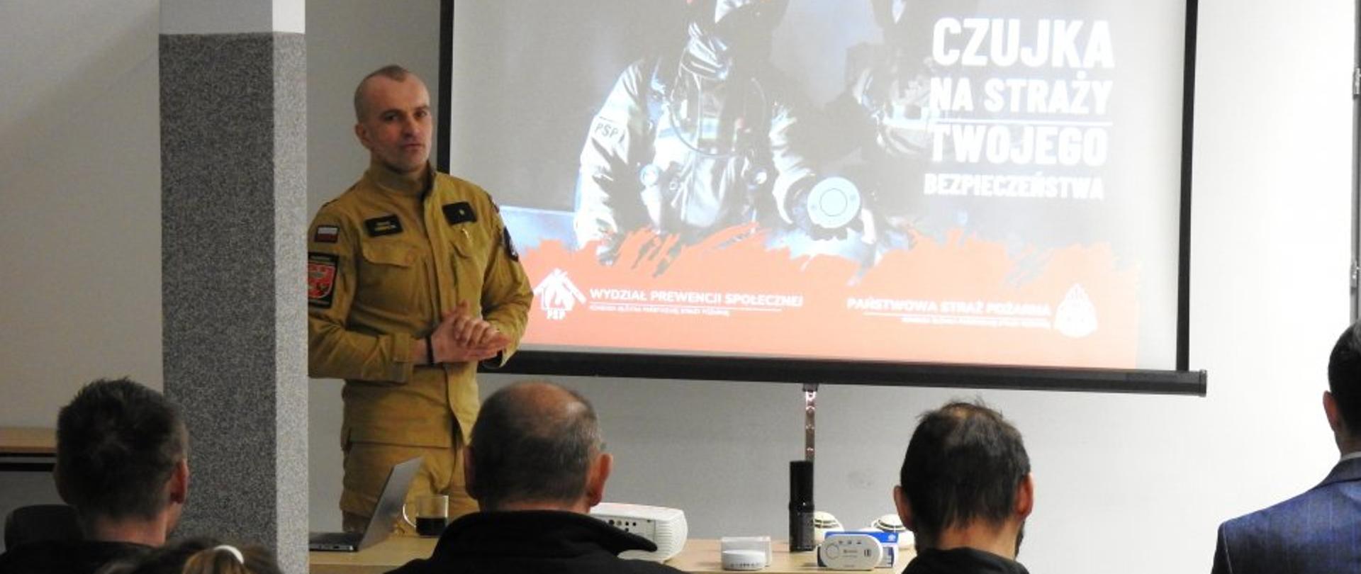 Strażak w piaskowym mundurze prowadzi prelekcję w sali szkoleniowej. Za nim znajduje się ekran z grafiką kampanii „Czujka na straży Twojego bezpieczeństwa”. Na pierwszym planie widać tyły głów słuchaczy, w tym osobę w czarnej bluzie z napisem „STRAŻ”. Na stole przed prowadzącym leżą rozłożone czujniki dymu i tlenku węgla.