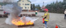 Pracownica zabezpieczona w hełm oraz kurtkę specjalną gasi płomienie przy użyciu gaśniczy