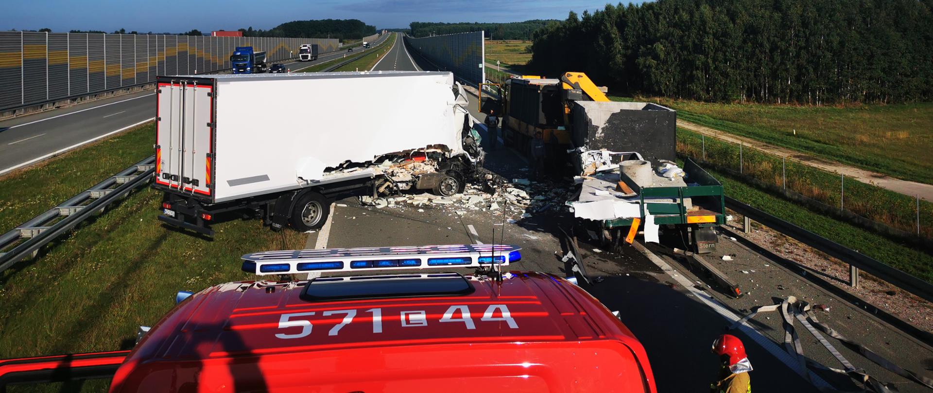 Zdjęcie przedstawia zablokowaną drogę ekspresową przez 2 samochody ciężarowe. Biały samochód ciężarowy po uderzeniu w przyczepę ustawioną na prawym poboczu drogi zatrzymał się w poprzek drogi. 