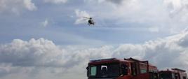 Zdjęcie przedstawia plac budowy, maszyny budowlane oraz pojazdy pożarnicze, które przyjechały do zdarzenia z poszkodowanym, którego poraził prąd elektryczny. Nad pojazdami pożarniczymi helikopter Lotniczego Pogotowia Ratunkowego przygotowującego się do lądowania.