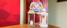 Na stole stoi symboliczny lampion z Światełkiem Pokoju. Zdjęcie wykonane w sali konferencyjnej strażnicy. Na drugim planie baner z czerwonym tłem, logiem Państwowej Straży Pożarnej i napisem "Komenda Powiatowa Państwowej Straży Pożarnej w Ostrzeszowie"
