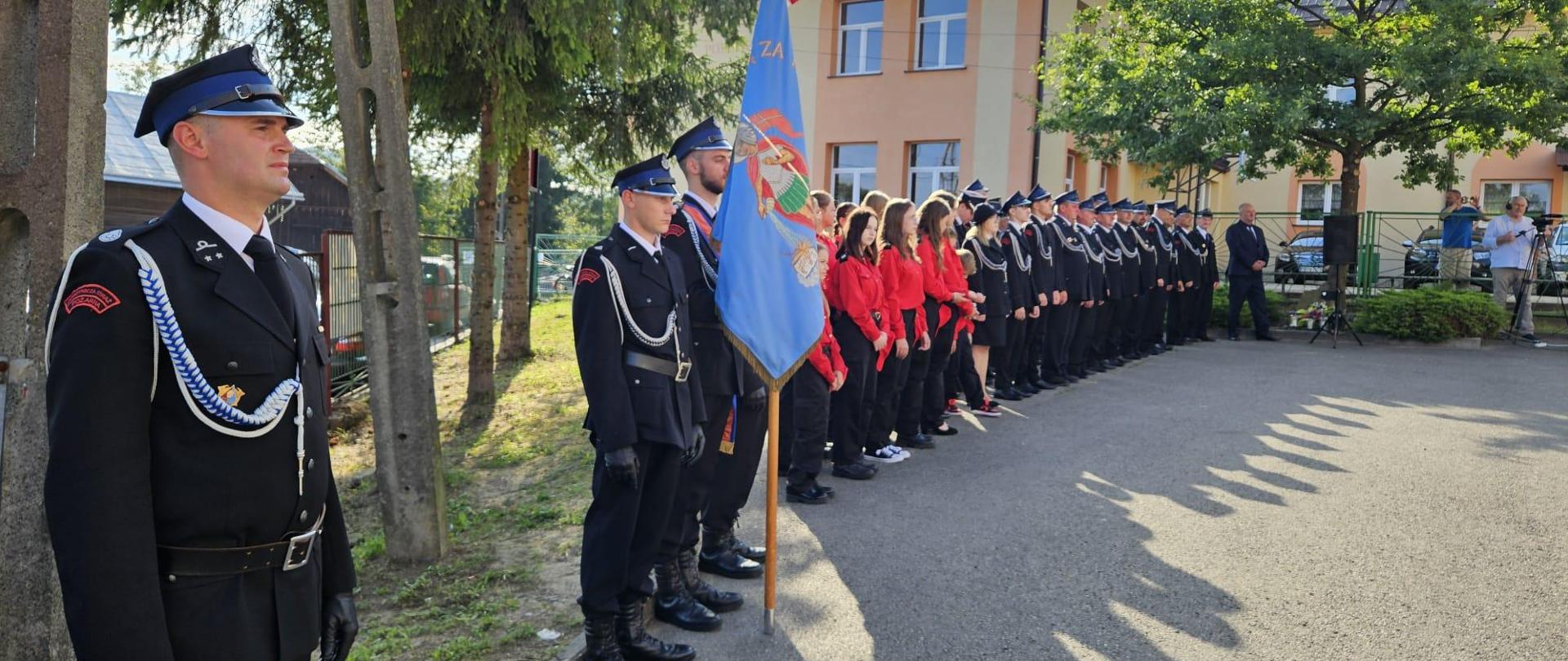 Kolorowa fotografia wykonana w pogodny dzień. Przedstawia strażaków w szyku zwartym z pocztem sztandarowym, młodzieżową drużyną pożarniczą i dowódcą uroczystości podczas 120 lecia na placu OSP w Domaradzu. W tle osoby cywilne.