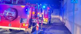 Ulica Sukiennicza w Rawiczu, na której stoja 2 wozy strażackie. Pierwszy z nich to samochód z podnośnikiem hydraulicznym. W koszu podnosnika znajduje się strażak. Z prawej strony ulicy widoczne sa ściany frontowe budynków. Pora nocna.