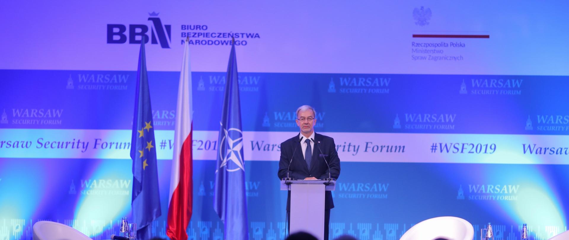 Minister Kwieciński stoi przy mównicy. Za nim stoi flaga UE, Polski i NATO oraz rozłożona jest ścianka na której znajdują się napisy Warsaw Security Forum, #WSF2019, Ministerstwo Spraw Zagranicznych i Biuro Bezpieczeństwa Narodowego. 