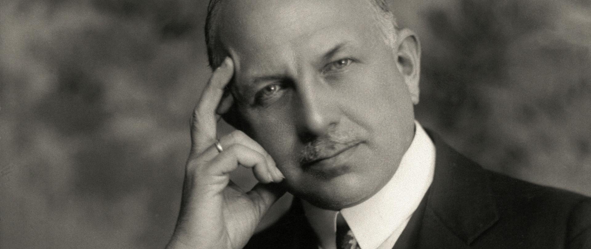 Karol Bertoni - portret mężczyzny podpierającego głowę ręką. Wąsy, siwe włosy, ciemna marynarka, musznik, ciemna kamizelka. Łokieć oparty na książce.