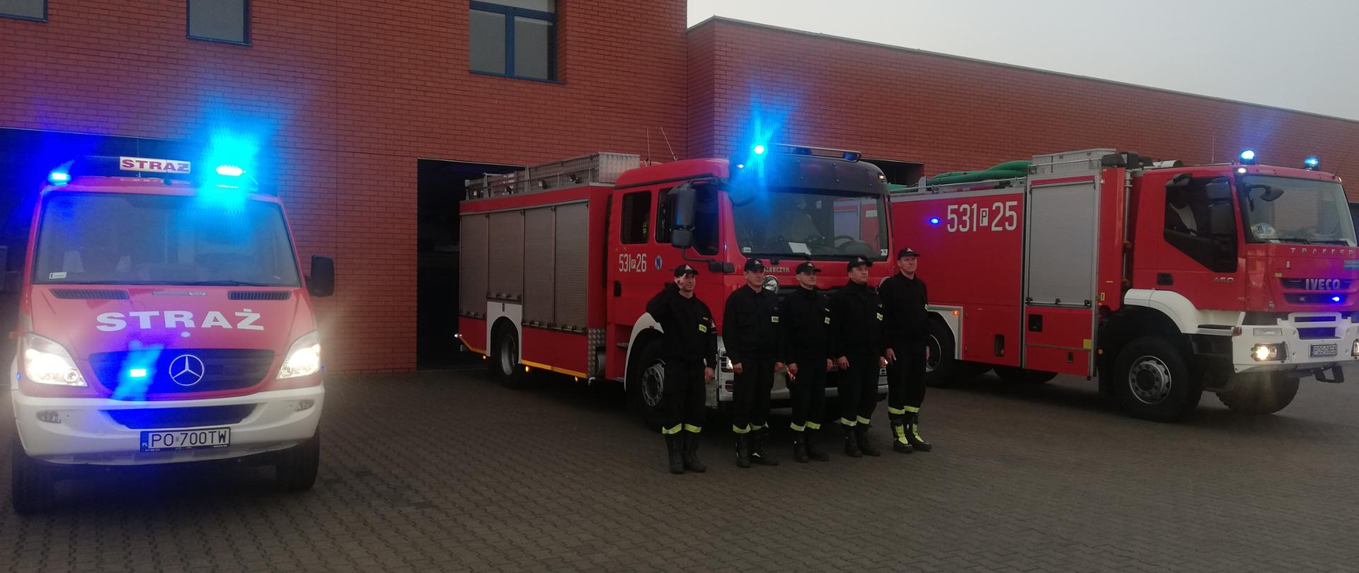 Pięciu strażaków oddaje hołd, w tle samochody strażackie z włączonymi światłami błyskowymi niebieskimi oraz komenda.