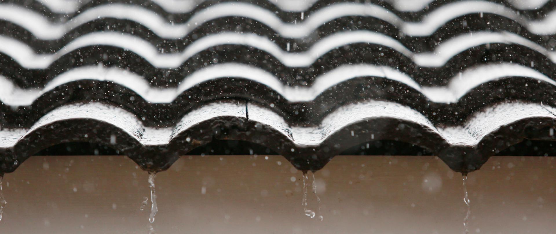 Na zdjęciu widać część falistego dachu z ciemnych dachówek, po którym spływa deszcz. 