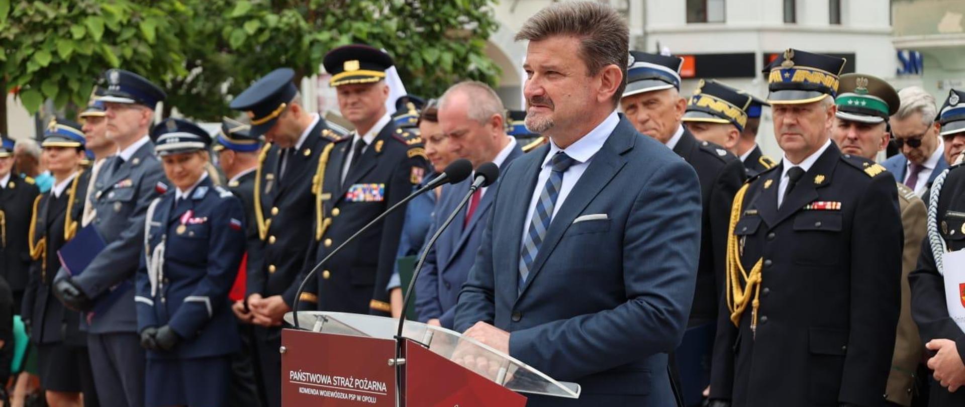 Wicewojewoda Opolski Piotr Pośpiech podczas przemówienia na Wojewódzkich Obchodach Dnia Strażaka w Krapkowicach. Na zdjęciu widać jak stoi przy czerwonej mównicy. Za nim można zobaczyć pozostałych uczestników uroczystości. 