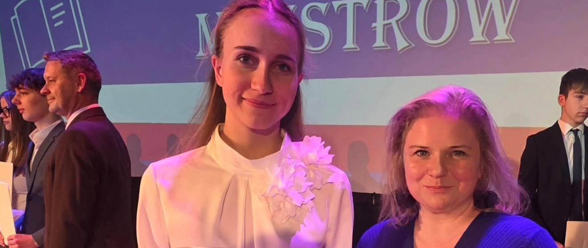 na zdjęciu w górnej części niebieski napis gala stypendiów prezesa rady ministrów, poniżej dwie kobiety o blond włosach trzymające bialy dyplom