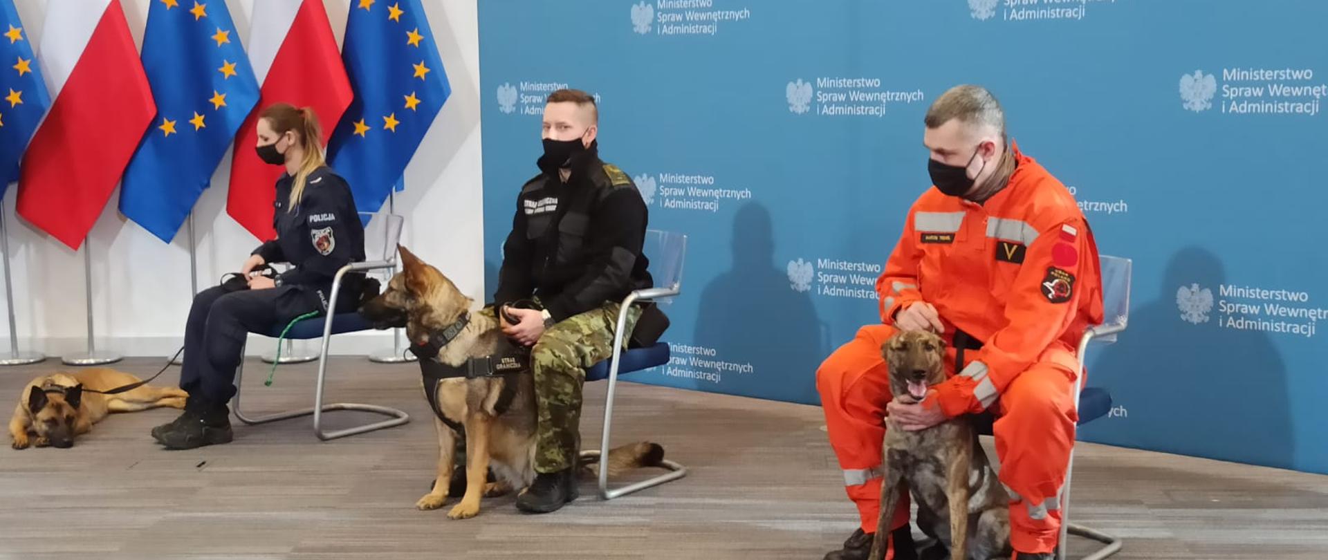 Na tle baneru MSWiA oraz flag Polski oraz Unii Europejskiej siedzi 3 funkcjonariuszy podległych MSWiA wraz z psami ratowniczymi.