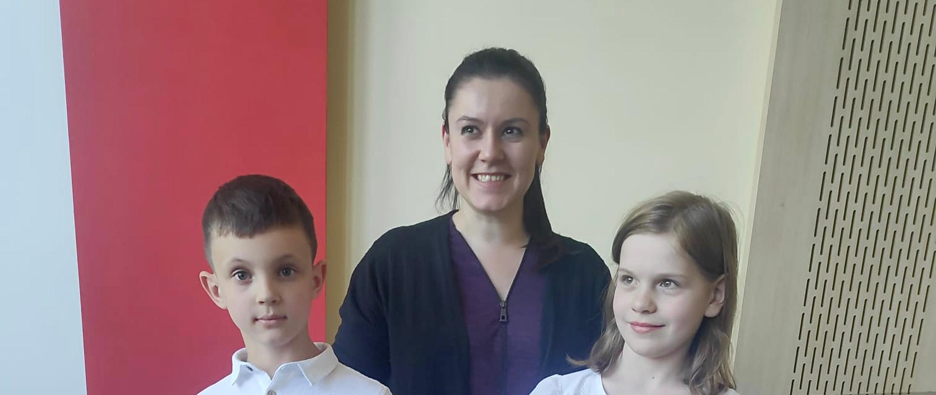 Chłopiec i dziewczynka trzymają dyplomy. Między nimi stoi kobieta