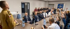 Zastępca Opolskiego Komendanta Wojewódzkiego Państwowej Straży Pożarnej wita gości - czeskich strażaków (w tym strażaczki) w świetlicy Ośrodka Szkolenia Komendy Wojewódzkiej Państwowej Straży Pożarnej w Opolu. Strażacy Republiki Czech siedzą przy dwóch długich stołach w mundurach wyjściowych. Komendant w umundurowaniu służbowym. W tle widoczny sztandar Szkoły Podoficerskiej w Opolu wiszący w gablocie.