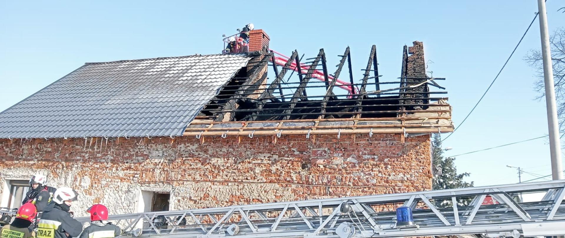Na zdjęciu widoczny budynek po pożarze poddasza i dachu. W dole zdjęcia widocznych czterech strażaków (2 OSP i 2 PSP) stojących przy opuszczonej drabinie mechanicznej. Dach budynku rozebrany w połowie. Odsłonięta część dachu z widoczną spaloną częścią więźby dachowej. W tle - za budynkiem - widoczny podnośnik hydrauliczny, strażacki w koszu, którego w okolicy komina pracuje dwóch strażaków.