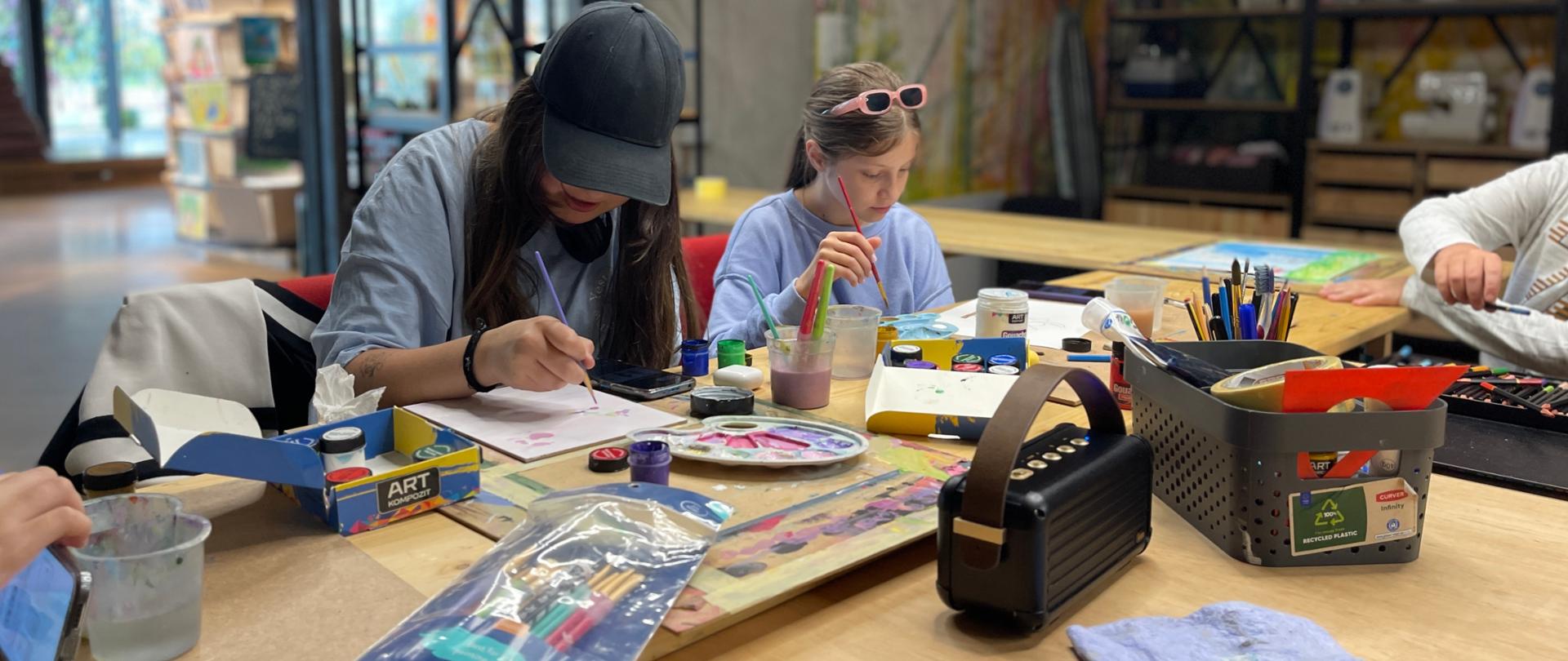 Przy stole siedzą uczestnicy zajęć i malują obrazki przy użyciu farb