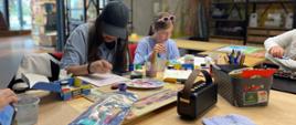 Przy stole siedzą uczestnicy zajęć i malują obrazki przy użyciu farb