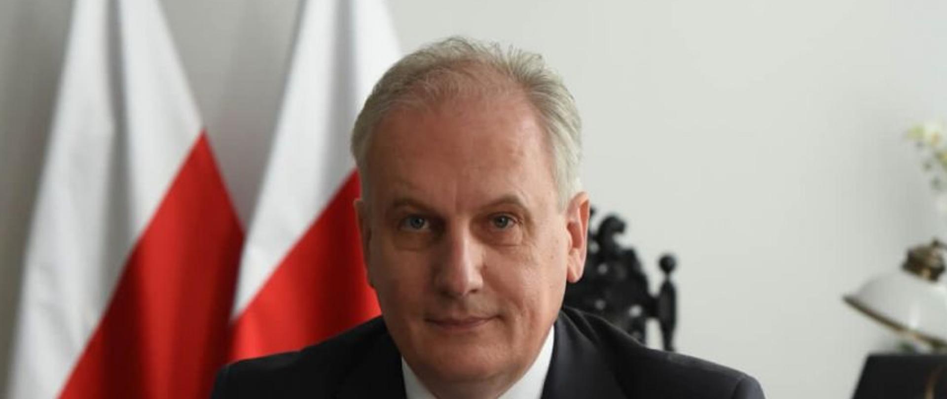 Wojewoda Pomorski Dariusz Drelich podpisujący dokumenty w gabinecie. Na biurku wojewody pomorskiego leżą kwiaty w barwach narodowych, W tle flagi państwowe i godło narodowe na ścianie.