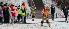 Strażak w pełnym umundurowaniu ochronnym, w kasku i kamizelce odblaskowej, przeciąga linę po zaśnieżonym terenie podczas pokazu dla dzieci. Po lewej stronie stoi grupa uczniów w zimowych kurtkach i czapkach, obserwujących demonstrację. W tle widać innych strażaków oraz schody prowadzące na wzgórze pokryte śniegiem.