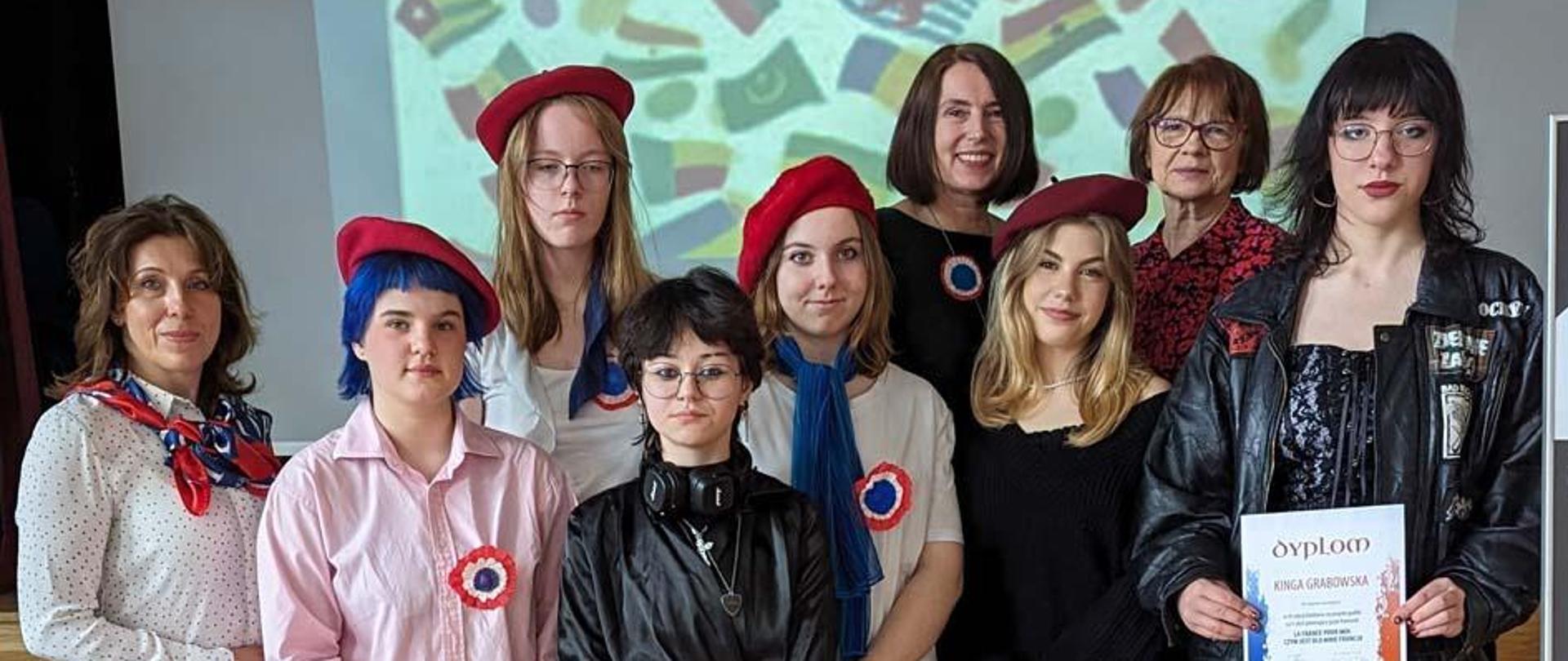 Trzy kobiety i między nimi sześć młodych osób. W tle wyświetlacz z wyświetlonym napisem La fête de la francophonie i kilkudziesięcioma flagami