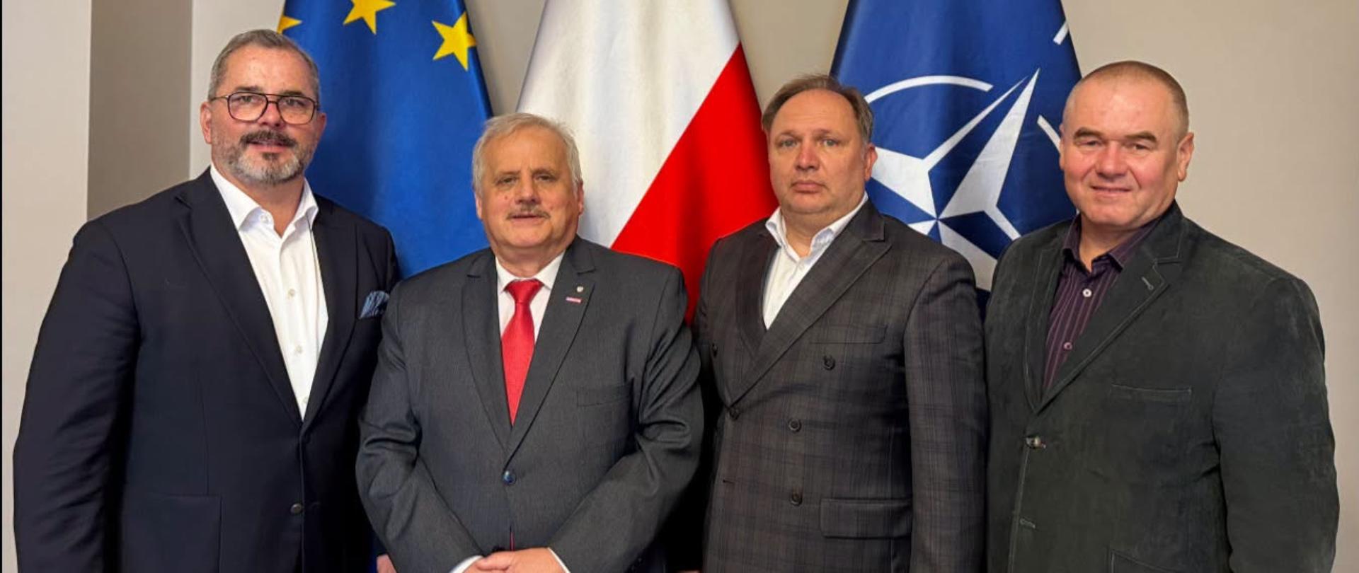 Grupa oficjeli w garniturach, w tym wicewojewoda, stoją na tle flag UE, Polski i NATO