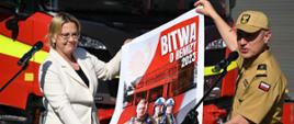 Komendant Główny PSP wraz z pani Minister Klimatu i środowiska trzymają rozciągnięty baner. Za nimi wóz pożarniczy. Komendant mówi do mikrofonu.