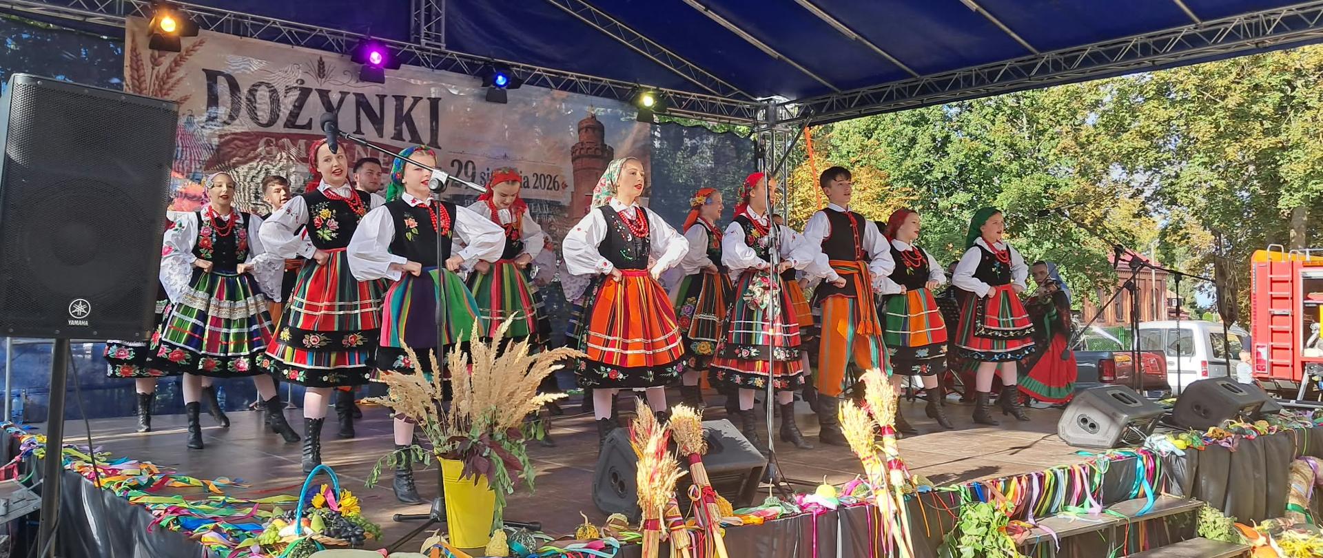 Scena podczas festynu, na której występuje zespół taneczny w bardzo kolorowych strojach.