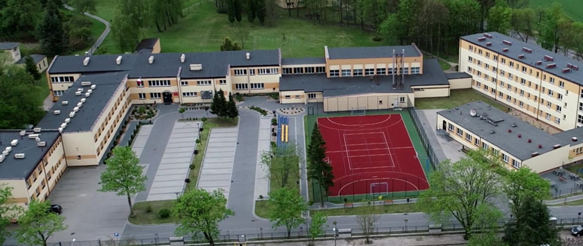 Widok z lotu ptaka na budynek główny z parkingiem, boiskiem i internatem, u góry park szkolny, okoliczne zabudowania i pola w zielonych i żółtych barwach