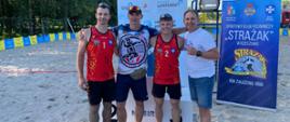 Czterech mężczyzn pozuje do wspólnego zdjęcia na piaskowym boisku do siatkówki plażowej. Dwóch z nich nosi czerwone stroje sportowe z numerami 1 i 2 oraz logo Sportowego Klubu Pożarniczego „Strażak” w Rzeszowie. Jeden mężczyzna ubrany jest w koszulkę z grafiką siatkarską, a ostatni w białą koszulkę i dżinsowe spodenki. W tle widoczny jest baner klubu „Strażak”, tablice sponsorów, boisko i parasol przeciwsłoneczny.