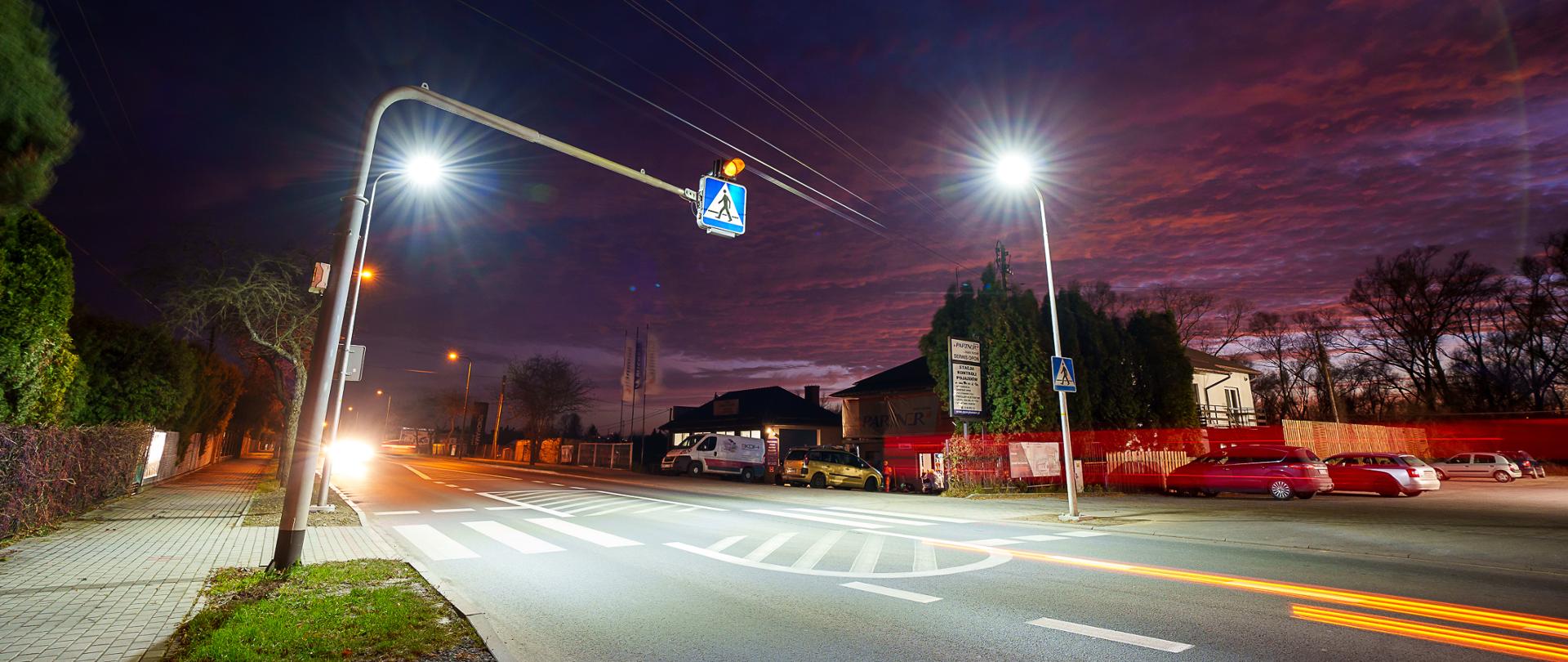 Na zdjęciu widać przejście dla pieszych przy drodze oświetlonej jasnymi lampami ulicznymi. Jest wczesny wieczór lub noc; niebo ma ciemne, fioletowo‑czerwone barwy. Po lewej stronie jedzie samochód, którego światła tworzą jasną smugę. Wzdłuż drogi stoją domy, zaparkowane auta i ogrodzenia. Nad przejściem umieszczono znak z podświetleniem.