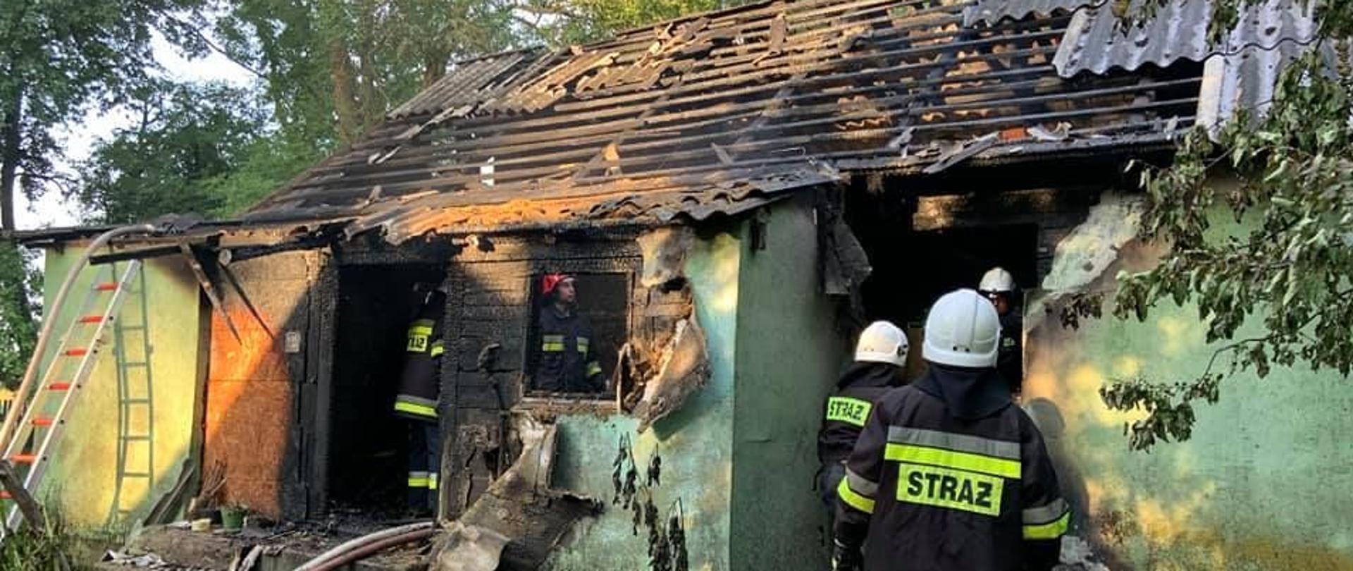 Na tle zielonego budynku z spalonym do połowy dachem, na pierwszym planie stoi tyłem dwóch strażaków w czarnych ubraniach specjalnych z białymi hełmami. wewnątrz budynku widać jeszcze trzech strażaków. W około budynku porozrzucane nadpalone elementy dachu.